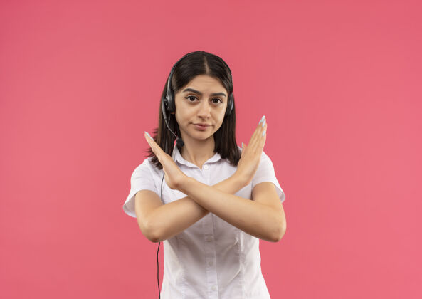 手势穿着白衬衫戴着耳机的年轻女孩 双手交叉站在粉红色的墙上做着停车标志女性女孩交叉