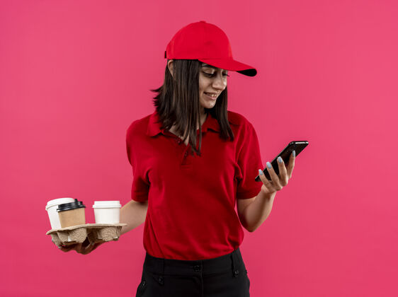 移动穿着红色马球衫 戴着帽子 拿着食品包的年轻送货女孩站在粉红色的墙上微笑着看着她的智能手机屏幕食品马球站着