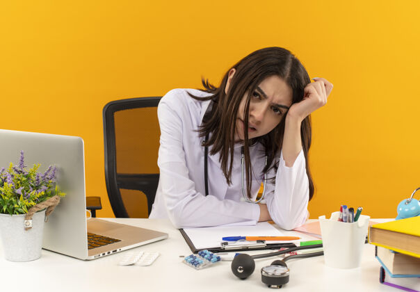 工作年轻的女医生穿着白大褂 拿着听诊器望着前方 困惑而焦急地坐在桌子旁 手提电脑和文件盖在橙色的墙上办公室坐着桌子