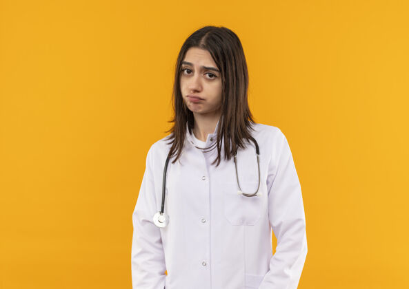 女人年轻的女医生穿着白大褂 脖子上戴着听诊器 眉头紧锁地站在橘色的墙上望着前方女人脸看