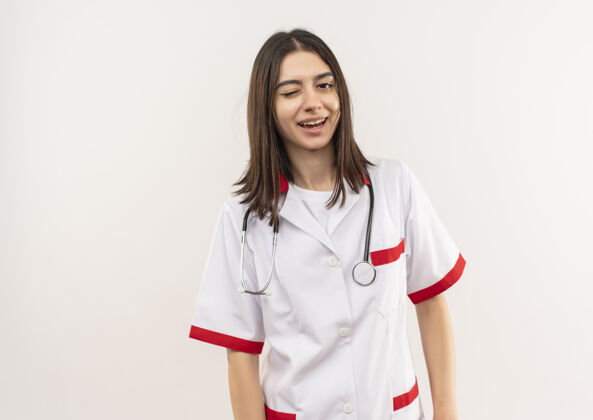看年轻的女医生穿着白大褂 脖子上戴着听诊器 微笑着眨着眼睛望着站在白墙上的前面人周围人