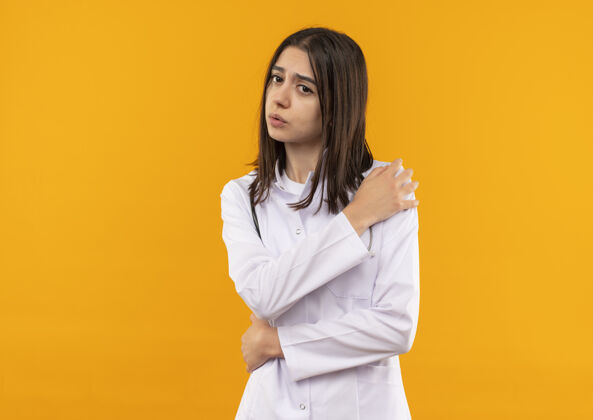 公民年轻的女医生穿着白大褂 脖子上戴着听诊器 身体不适 摸着肩膀 站在橘色的墙上疼医生周围姿势
