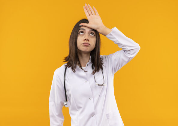 外套年轻的女医生穿着白大褂 脖子上戴着听诊器 站在橙色的墙上 头上挂着一个失败者的牌子 看上去很困惑头周围姿势