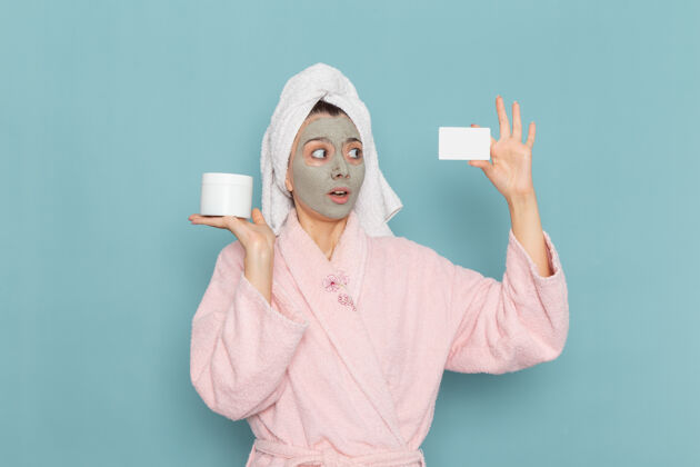 面部正面图：年轻女性 穿着粉色浴袍 淋浴后 脸上戴着口罩 手拿蓝色墙壁上的卡片 淋浴清洁美容自护霜后实验服浴袍