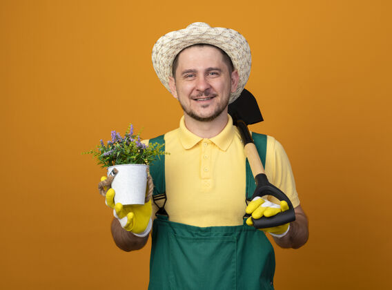 微笑年轻的园丁穿着连体衣 戴着帽子 拿着铲子和盆栽植物 站在橙色的墙上 面带微笑地看着前方市民人连身衣