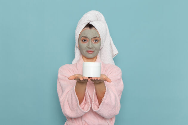 正面正面图年轻女性穿着粉色浴衣 脸上戴着口罩拿着面霜可以在蓝色墙上淋浴清洗美容自护霜成人浴袍面霜