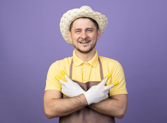 工作年轻的园丁戴着工作手套 穿着连体衣 戴着帽子 双手交叉 站在紫色的墙上 露出v字标志 微笑着人人年轻人