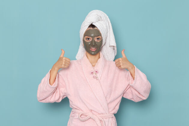 人正面图：年轻女性 穿着粉色浴袍 淋浴后 脸上戴着深色面罩 蓝色桌上 美丽的水霜 自我护理淋浴浴室视野年轻人浴袍
