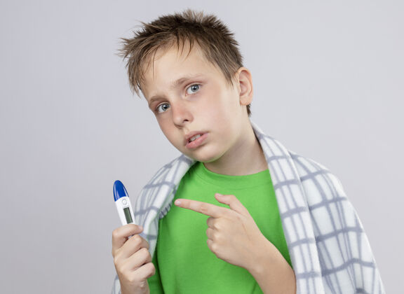 包着生病的小男孩 穿着绿色t恤 裹着毯子 身体不适 拿着温度计 手指指着它 站在白墙上 心烦意乱不舒服男孩小