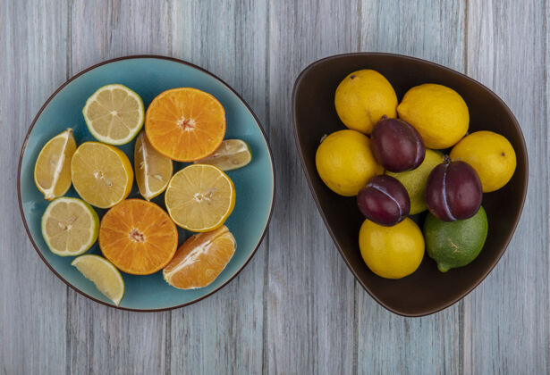 柠檬顶视图李子在一个碗与柠檬楔石灰和橙色在一个盘子上的灰色背景李子视野盘子