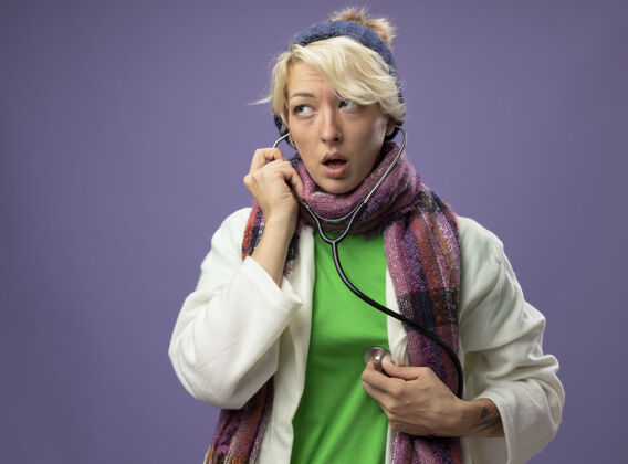 周围生病的不健康的女人 短发 戴着暖和的围巾 戴着帽子 脖子上戴着听诊器 站在紫色的墙上听着自己的心跳女人短裤生病