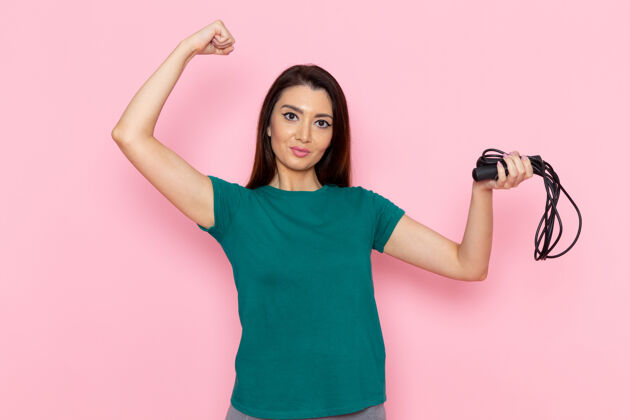 锻炼正面图身着绿色t恤的年轻女性手持跳绳 在浅粉色墙壁上弯曲腰部运动锻炼美丽苗条的女运动员运动观点前面