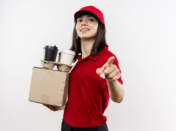 帽子年轻的送货员穿着红色制服 戴着帽子 拿着盒装和咖啡杯 用食指指着一张笑脸站在白色的墙上盒子站着穿