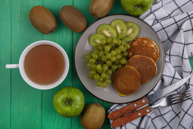 茶顶视图一杯茶与猕猴桃绿色葡萄和薄煎饼在一个盘子上与刀叉在一个格子毛巾在绿色的背景刀盘子上衣