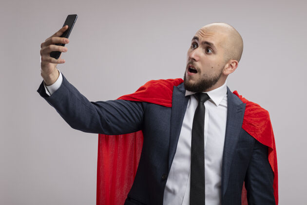 男人超级英雄胡子商人在红斗篷使用智能手机自拍被困惑站在白墙自拍使用超级英雄
