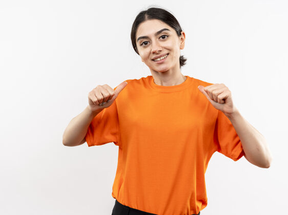 年轻穿着橙色t恤的年轻女孩站在白色的墙上 指着自己微笑着 自信 快乐 积极她自己站着积极