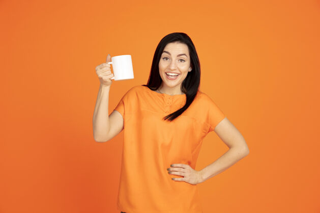 人类橙色工作室背景上的白种人年轻女子肖像穿着衬衫的漂亮黑发女性模特人类情感的概念 面部表情 销售 广告广告空间喝咖啡或茶女性员工肖像