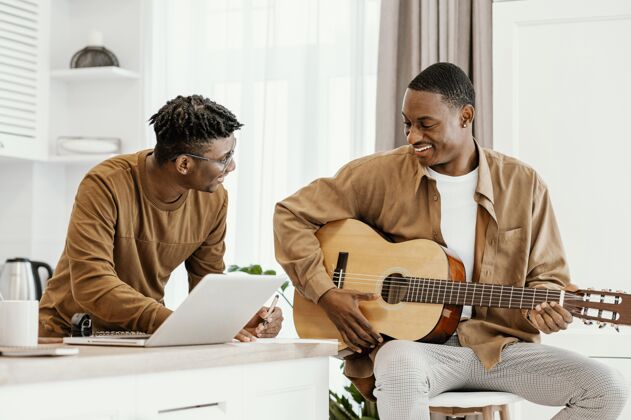 人才笑脸男音乐家在家的前视图在椅子上弹吉他和使用笔记本电脑表演男人音乐家