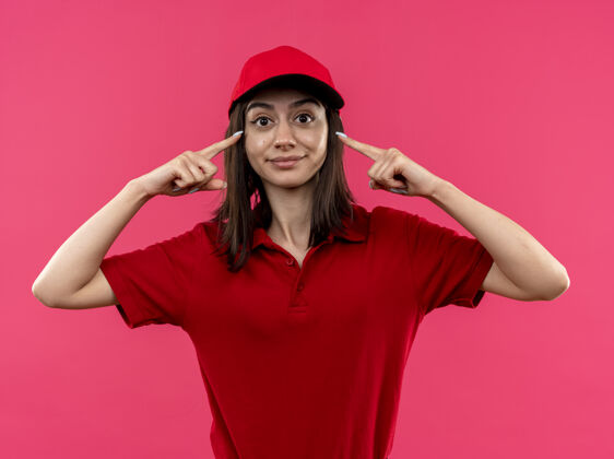 看身穿红色马球衫 头戴帽子的年轻送货女孩站在粉红色的墙上 面带微笑地指着自己的太阳穴 专注于一项任务任务微笑马球