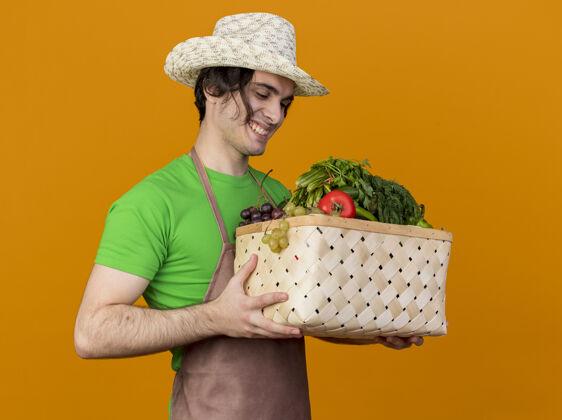 帽子年轻的园丁 围着围裙 戴着帽子 手里拿着装满蔬菜的箱子 站在橙色的墙上 面带微笑地看着它蔬菜脸站着