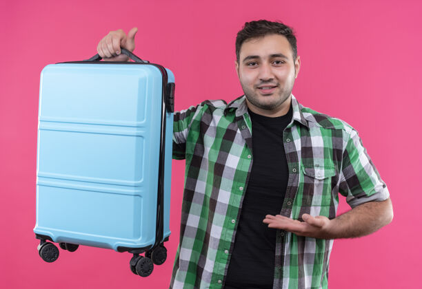 介绍穿着格子衬衫的年轻旅行家站在粉红色的墙上 手上带着微笑 自信地展示着他的手提箱男人姿势人