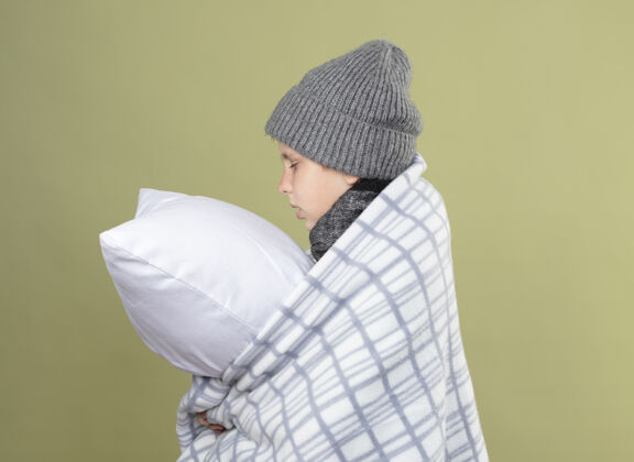 疾病生病的小男孩 戴着暖和的帽子 裹着毯子 抱着枕头 身体不舒服地站在轻墙上小温暖枕头