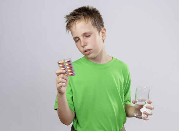 流感生病的小男孩穿着绿色t恤 站在白色的墙上 手里拿着一杯水和感冒药 感觉不舒服玻璃站着抱着