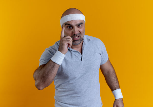 运动员戴着头巾的成熟运动型男子望着前面 用食指警告 严肃的脸站在橙色的墙上肌肉人警告