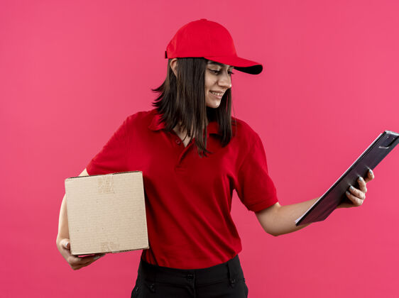 举行穿着红色马球衫和帽子的年轻送货女孩拿着盒子和剪贴板看着它微笑着 幸福的脸站在粉红色的墙上脸包装站