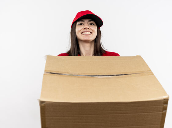 女孩年轻的送货员身穿红色制服 戴着帽子 手里拿着大纸盒 脸上带着微笑站在白色的墙上穿制服大