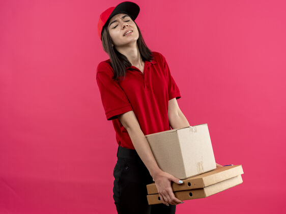 盒子年轻的送货女孩穿着红色马球衫 戴着帽子 手里拿着沉重的盒装 站在粉色的墙上显得疲惫和紧张累了送货紧张