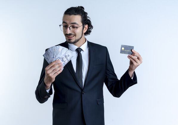 生意一个穿着黑色西装戴着眼镜的商人站在白色的墙上 面带笑容地展示着现金和信用卡优雅脸男人