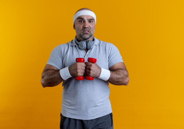 头带成熟的运动型男子戴着头巾 站在橙色的墙2上练习哑铃 看起来紧张而自信人运动员运动