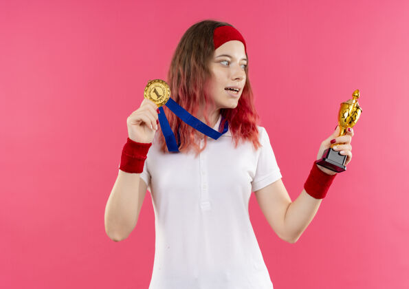 女性戴着头巾 脖子上戴着金牌 手拿奖杯 高兴地看着奖杯的年轻运动女性站在粉红色的墙上 走了出来运动看姿势
