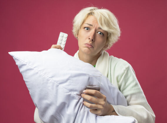 枕头生病的不健康的短发女人抱着枕头不舒服拿着一杯水和药站在粉红色的墙上看起来很沮丧抱着心烦药品