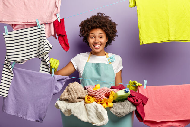 洗涤剂面带微笑的黑皮肤管家把干净的衣服和衣夹挂在洗衣线上洗家庭作业干