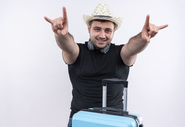 人穿着黑色t恤 戴着夏令帽 手拿手提箱的年轻旅行家站在白色的墙上 手指上挂着摇滚乐的符号手指夏天旅行