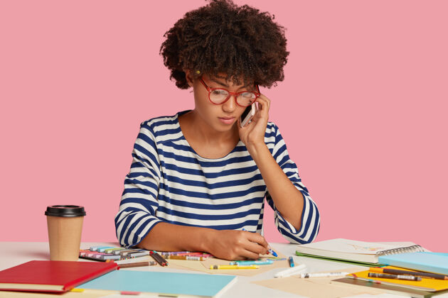 外观严肃忙碌的黑人女工人的照片 为项目工作制作插图 通过手机与合作伙伴交谈 戴着透明眼镜和条纹衣服 隔着粉红色的墙咖啡工人学习