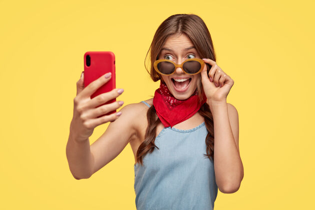 快乐喜气洋洋的黑发女人 喜欢为夏天买时髦的墨镜 准备度假 在手机上拍照 黄色墙壁上的模特女孩自拍魅力电话满意