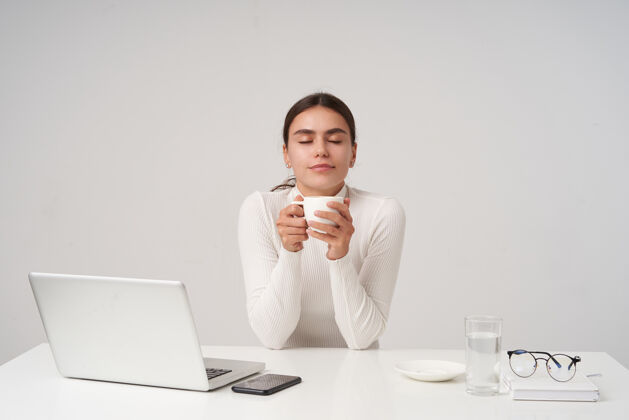 发型喜欢年轻漂亮的黑发女人 扎着马尾辫 喝着咖啡休息 拿着白色陶瓷杯 闭着眼睛愉快地微笑 坐在白色的墙上女士眼镜笔记本电脑