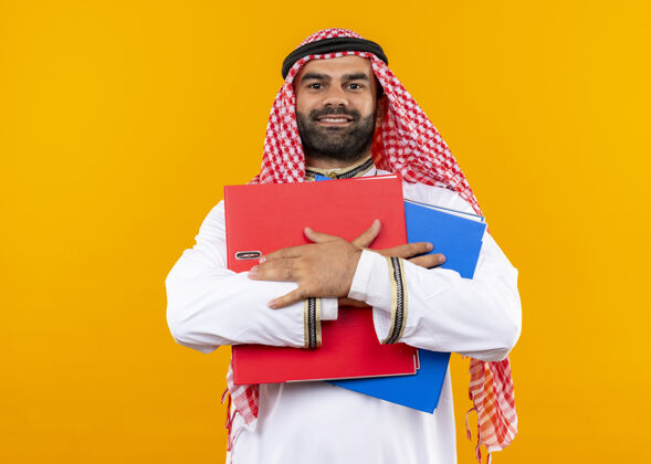 穿着身着传统服装的阿拉伯商人手持两个文件夹 微笑自信地站在橙色的墙上公民商人持有