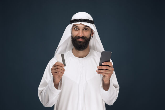 支付深蓝色工作室背景上的阿拉伯沙特商人肖像男子使用智能手机付账 网上购物或投注商业概念 金融 面部表情 人类情感面部男人民族