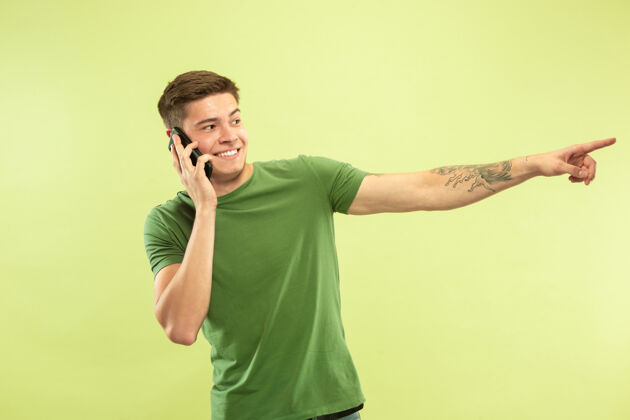 手势绿色工作室背景上的白人年轻人半身肖像穿着衬衫的漂亮男模人类情感的概念 面部表情 销售 广告打电话 指着 微笑专业公司衬衫