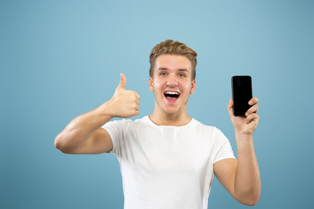 衬衫在蓝色工作室背景上的白人年轻人半身肖像穿着衬衫的漂亮男模人类情感的概念 面部表情 销售 广告显示电话屏幕 付款 投注面部商人表情