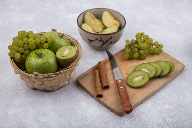 篮子侧视图猕猴桃片上有葡萄 肉桂和一把刀放在砧板上 绿色的苹果放在篮子里 切片放在碗里 背景是白色的肉桂碗壁板