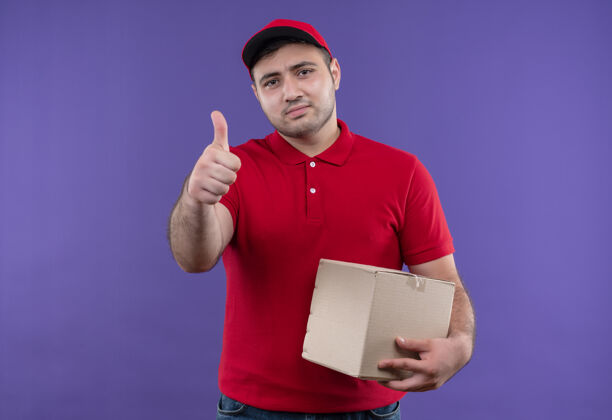盒子年轻的送货员穿着红色制服 戴着帽子 拿着盒子 微笑着 自信地竖起大拇指站在紫色的墙上站着年轻人男人