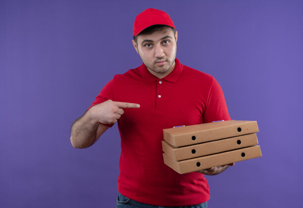 人年轻的送货员身穿红色制服 戴着帽子 拿着披萨盒 手指指着他们 站在紫色的墙上 看上去很自信看拿着年轻人