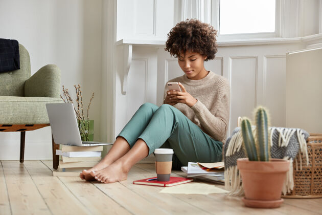 学生美丽的非洲女孩坐在地板上舒适的房间里 在网络上浏览个人资料 喝咖啡 与文学和笔记本电脑一起工作 在手机上聊天 穿着休闲毛衣和裤子扶手椅民族笔记本