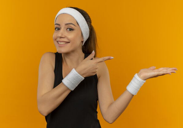 头带身穿运动服的年轻健身女 头戴头巾 快乐而积极地用胳膊和手指指着站在橙色墙上的一边运动运动装壁板