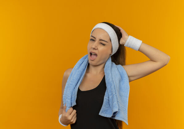 毛巾站在橘色的墙上 一个穿着运动服 头上戴着头巾 脖子上挂着毛巾的年轻健身女士看起来很困惑健康运动装站